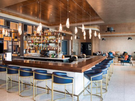 A modern bar interior with stylish seating, a well-stocked bar, pendant lighting, and contemporary decor in a spacious setting.