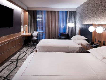 A modern hotel room with two neatly made beds, a flat-screen TV, a desk with a chair, and a window with curtains allowing minimal light.