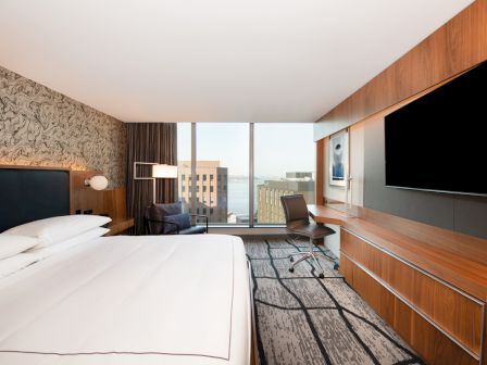 A modern hotel room with a large bed, wall-mounted TV, desk, and floor-to-ceiling windows offering a cityscape view.