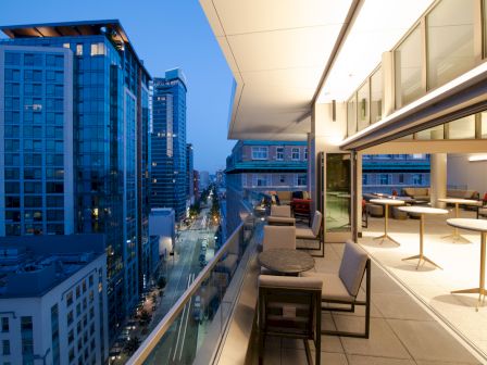 A modern balcony with tables and chairs overlooks a cityscape with tall buildings at dusk. The view captures an urban street below.