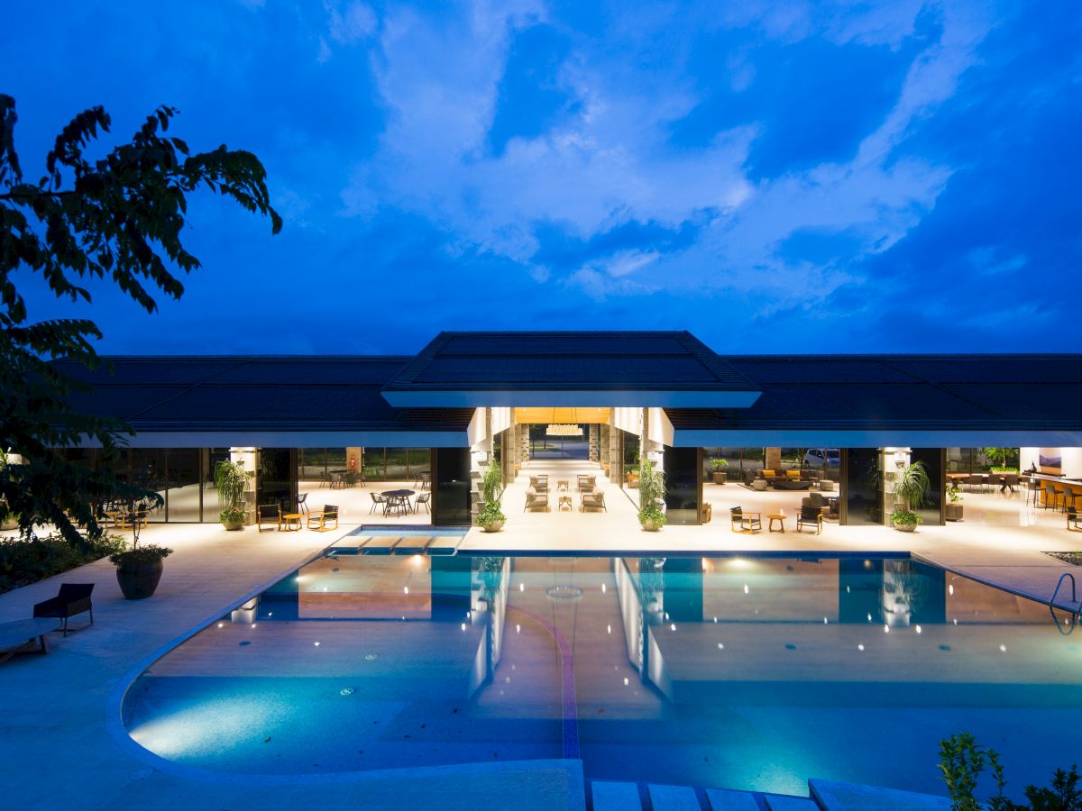 A modern building with an illuminated pool in the foreground, lounge areas inside the open structure, and a blue evening sky in the background.