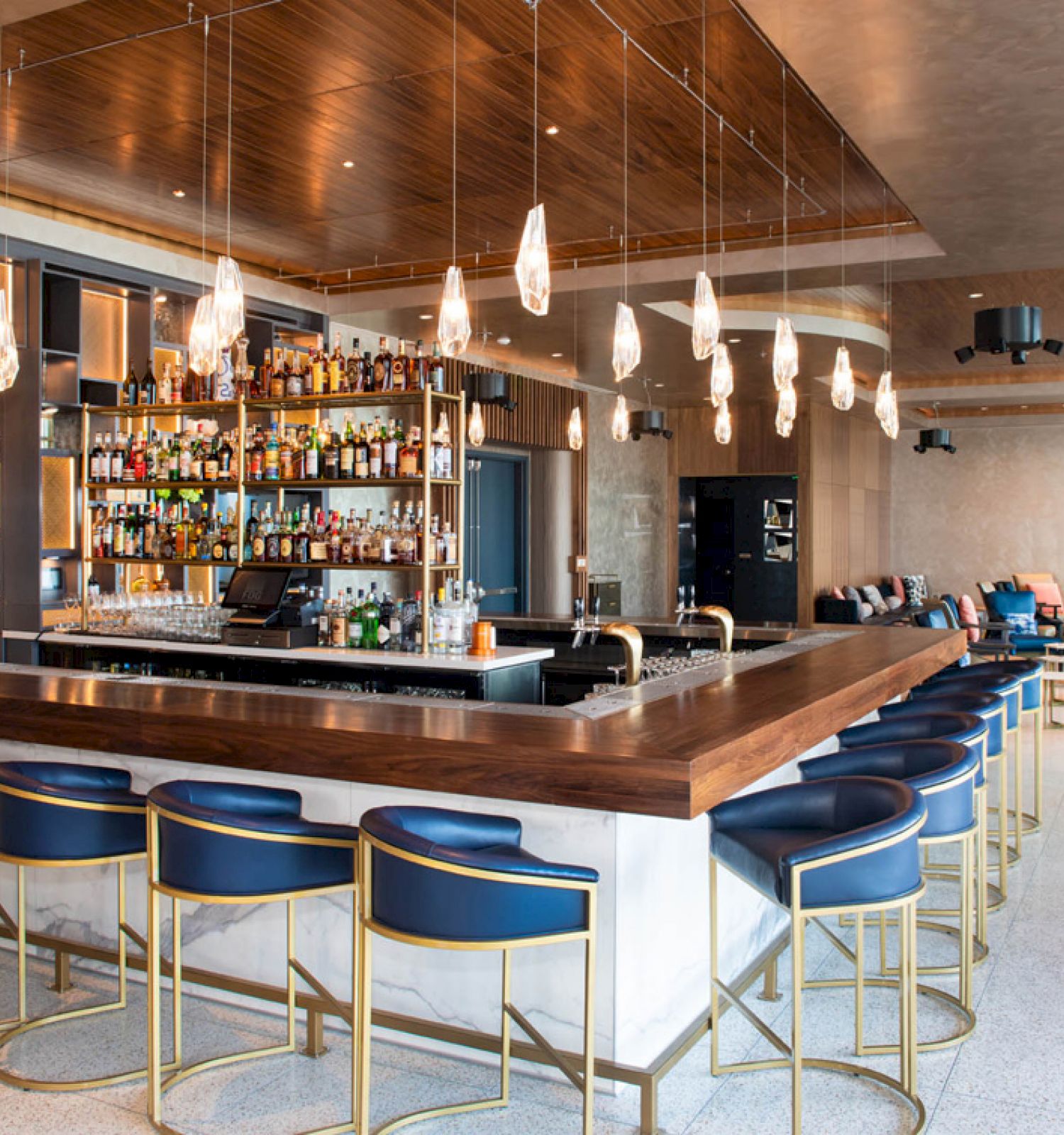 A modern bar with a wooden counter, blue stools, hanging lights, and a well-stocked back bar; cozy seating areas in the background.