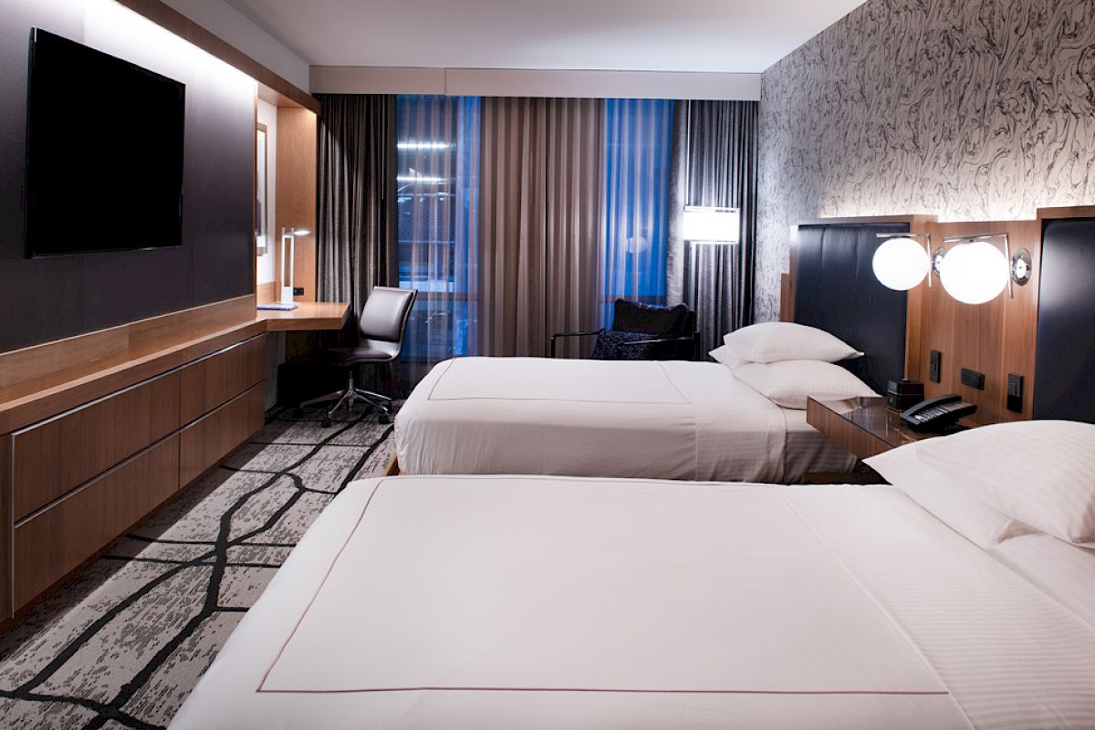 The image shows a modern hotel room with two neatly made beds, a large TV, a desk with a chair, and stylish decor.