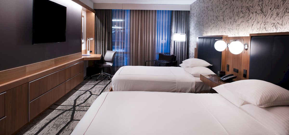 The image shows a modern hotel room with two neatly made beds, a large TV, a desk with a chair, and stylish decor.