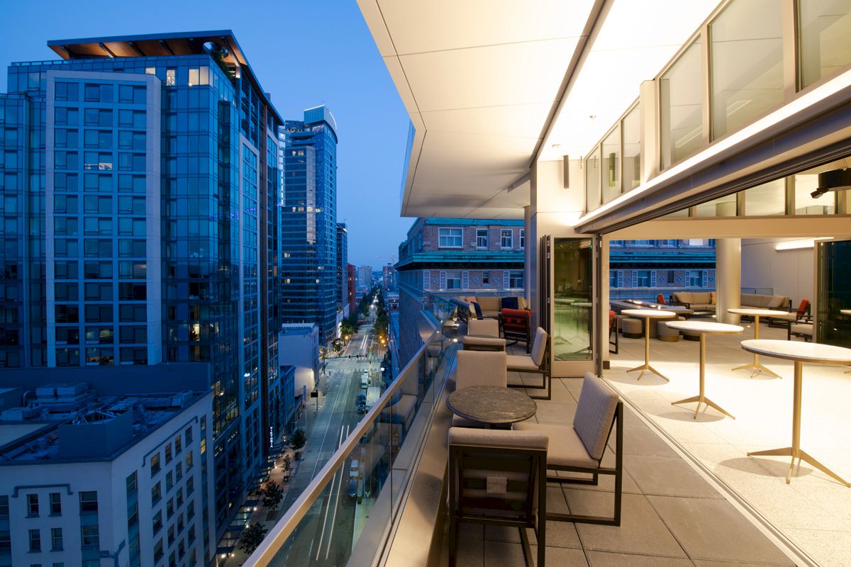 Modern cityscape with tall buildings and a sleek balcony featuring tables and chairs overlooking a vibrant street at dusk.