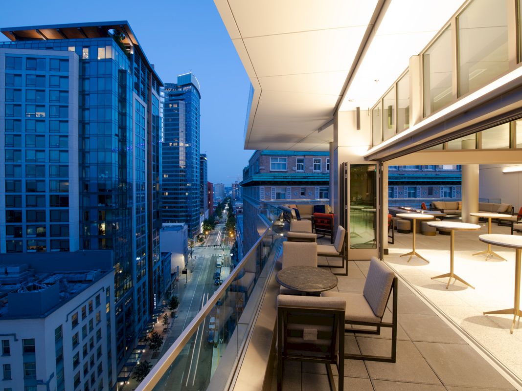 Modern cityscape with tall buildings and a sleek balcony featuring tables and chairs overlooking a vibrant street at dusk.