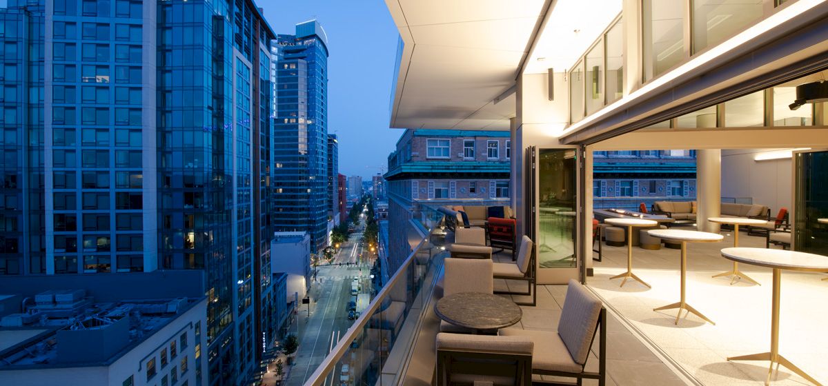 Modern cityscape with tall buildings and a sleek balcony featuring tables and chairs overlooking a vibrant street at dusk.
