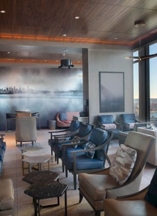 A modern, comfortably furnished lounge with ample seating and a view of a waterfront and skyline through large windows is shown.