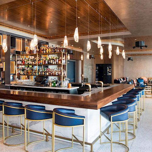 The image shows a modern and stylish bar with a wooden countertop, blue cushioned bar stools, hanging lights, and a full wall of liquor bottles.