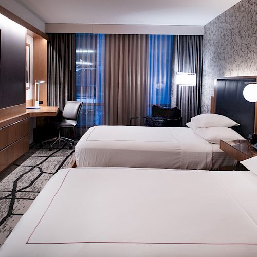 The image shows a modern hotel room with two double beds, a flat-screen TV, a work desk, and contemporary decor, lit by soft ambient lighting.