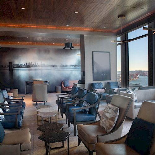 A modern lounge with comfortable seating, large windows showing a waterfront view, and a wall mural depicting a city skyline completes the scene.