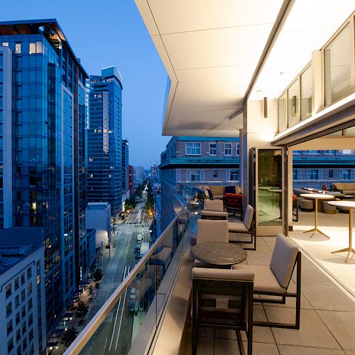 A modern balcony overlooking a cityscape with high-rise buildings, furnished with tables and chairs, and illuminated ambient lighting.