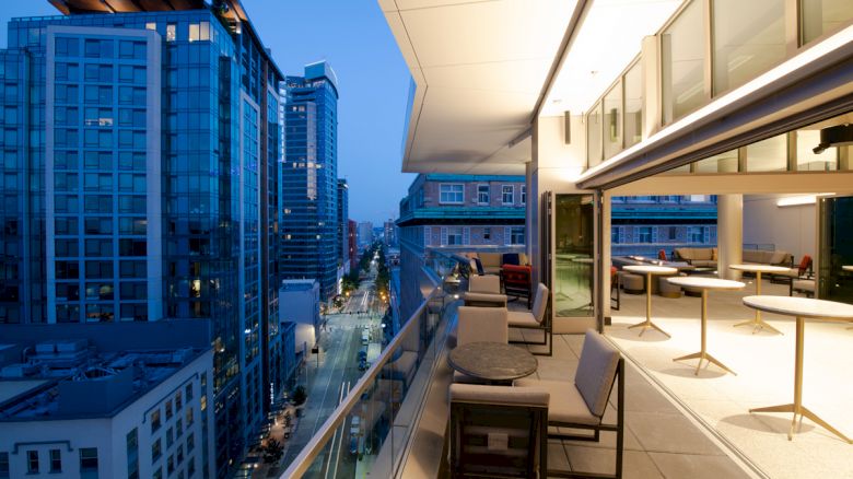 The image shows a modern cityscape at dusk with tall buildings. In the foreground, there is an outdoor terrace with tables and chairs.