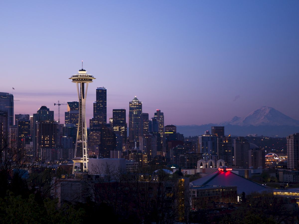 The image shows the Seattle skyline at dusk, with the iconic Space Needle prominently visible and Mount Rainier in the background, under a clear sky.