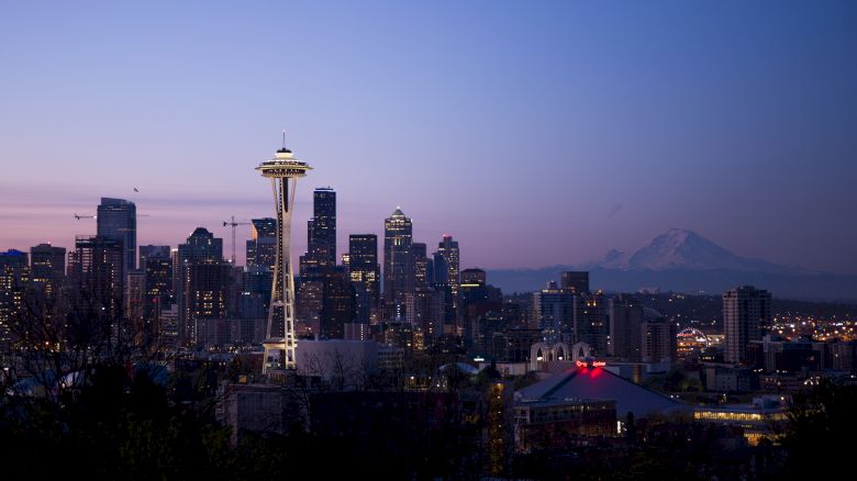 The image shows the Seattle skyline at dusk, with the iconic Space Needle prominently visible and Mount Rainier in the background, under a clear sky.