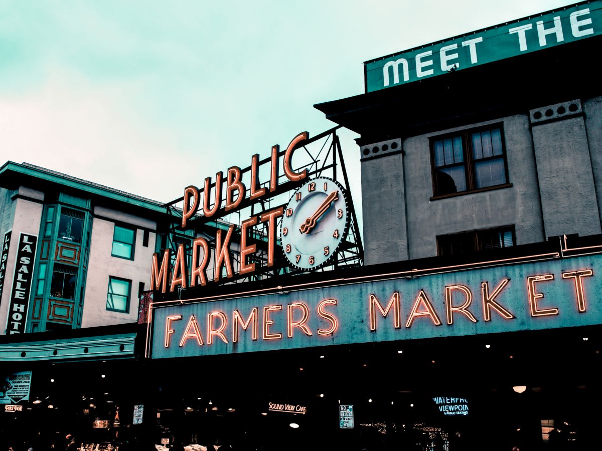 The image shows a neon sign for a "Public Market" and "Farmers Market" with a clock beneath it, surrounded by buildings.