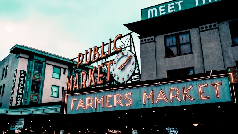 The image shows a neon sign for a "Public Market" and "Farmers Market" with a clock beneath it, surrounded by buildings.