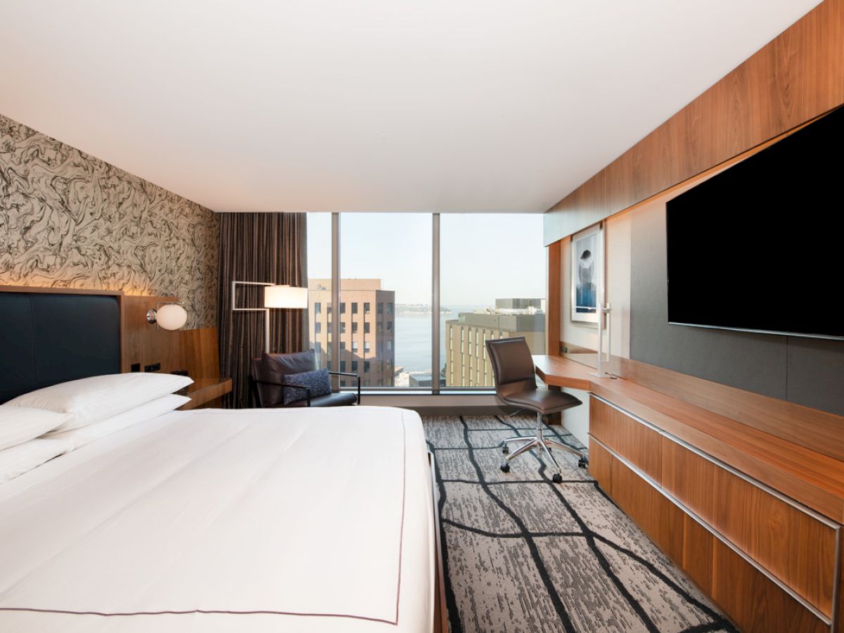 A modern hotel room features a large bed, wall-mounted TV, desk with chair, and a wide window with a city view.