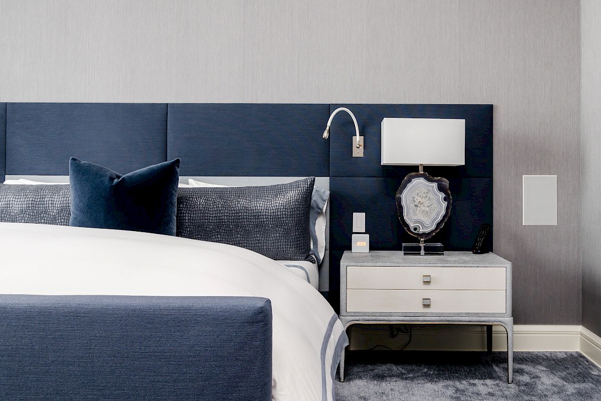 A modern bedroom with a blue and white color scheme, featuring a bed, two pillows, a bedside table with a lamp, and a wall-mounted reading light.