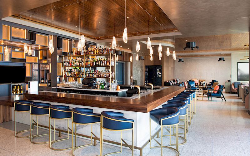 The image shows a modern bar with a sleek counter, blue bar stools, hanging light fixtures, and a well-stocked shelf behind the bar.