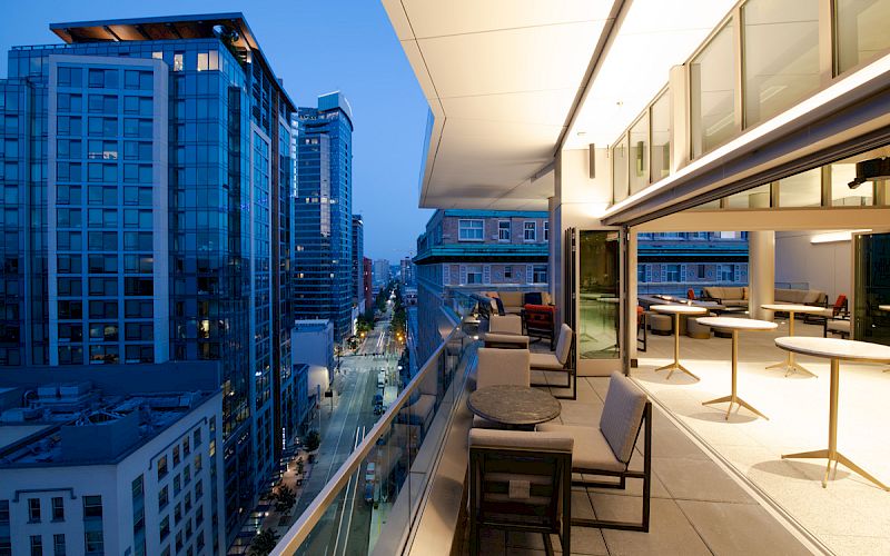 A modern balcony with seating and tables, overlooking a cityscape with tall buildings illuminated in the evening.