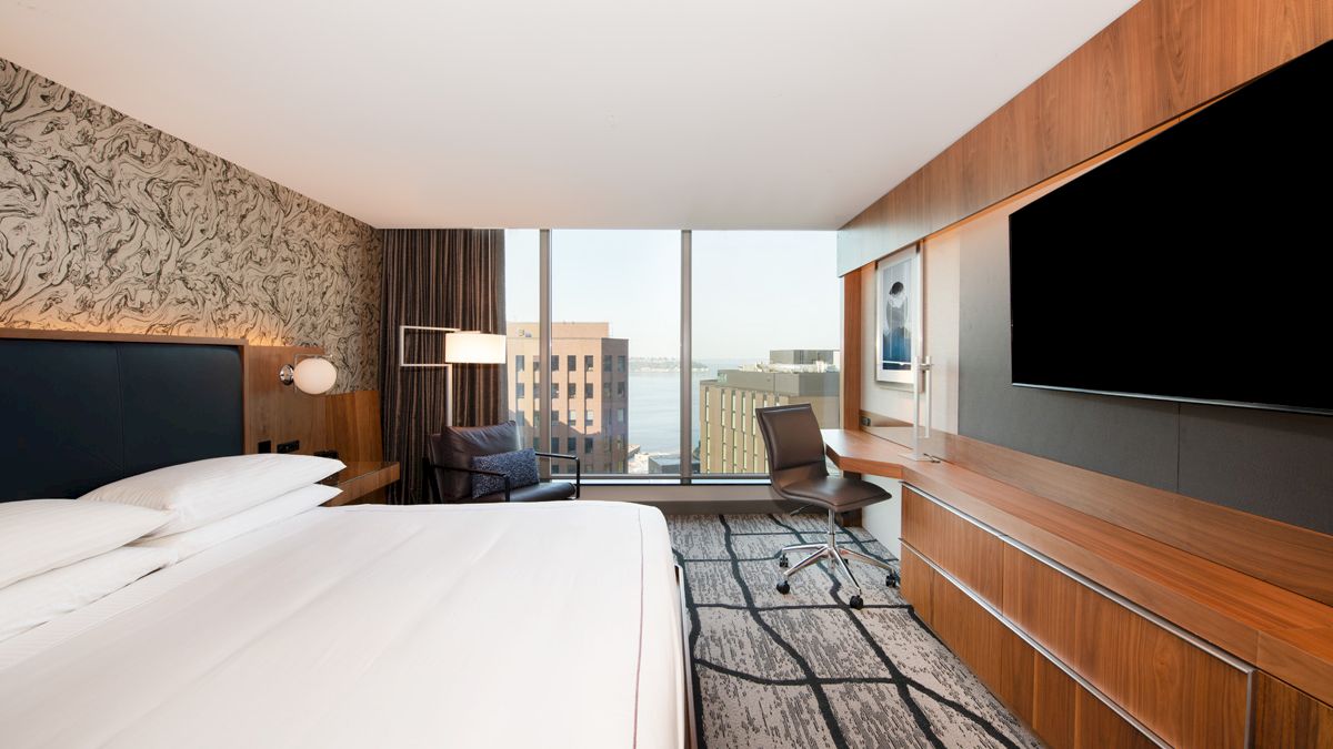 A modern hotel room features a king-sized bed, large flat-screen TV, desk area, and floor-to-ceiling windows with a city and water view.