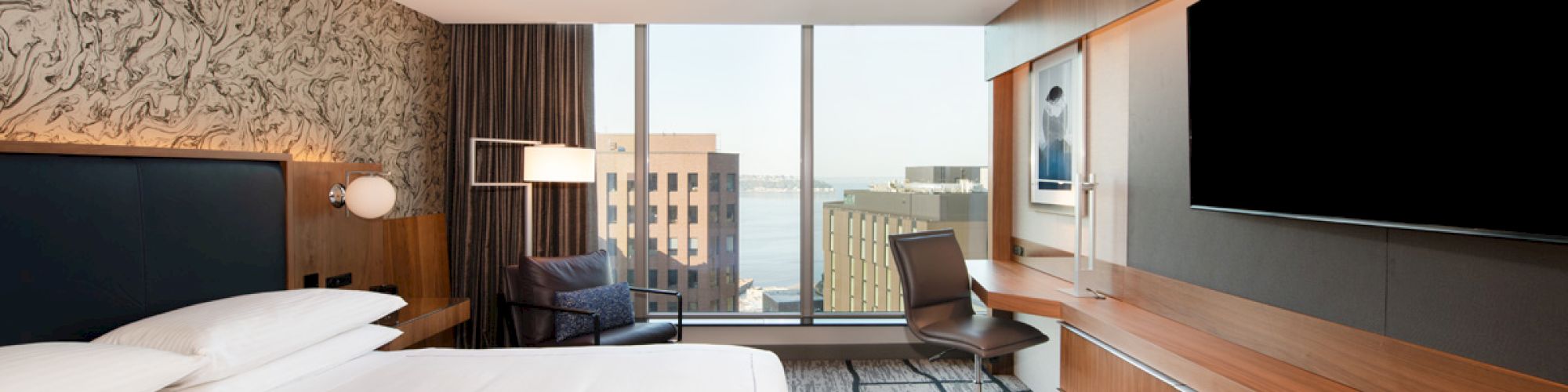 A modern hotel room features a king-sized bed, large flat-screen TV, desk area, and floor-to-ceiling windows with a city and water view.